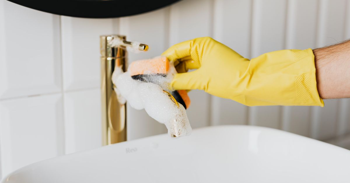 Une main gantée de jaune nettoie un robinet doré avec une éponge savonneuse au-dessus d'une baignoire ou vasque blanche.