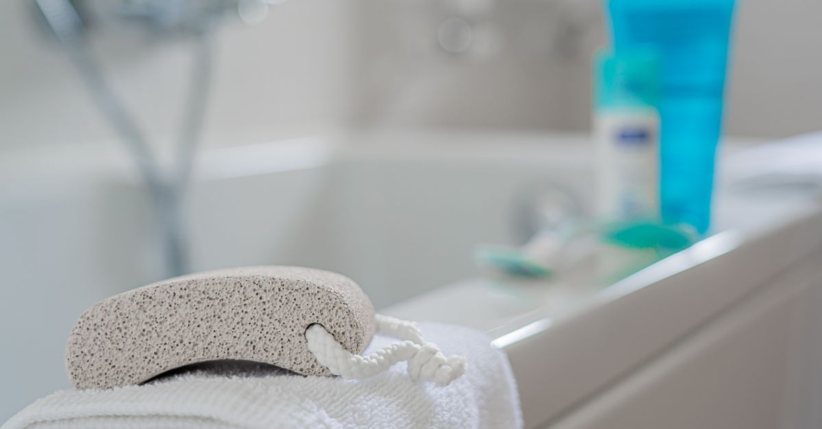 Une pierre ponce posée sur une serviette blanche au bord d'une baignoire, avec des flacons de produits en arrière-plan flou.