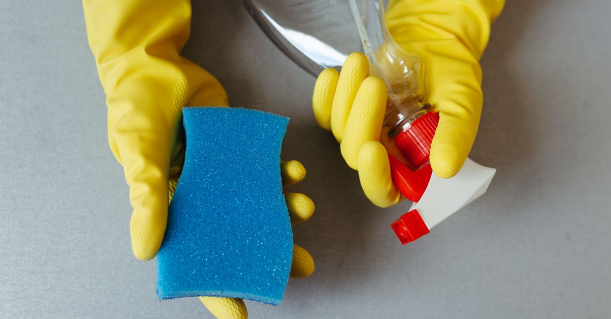 Des mains gantées de jaune tiennent une éponge bleue et un flacon de détergent ouvert.