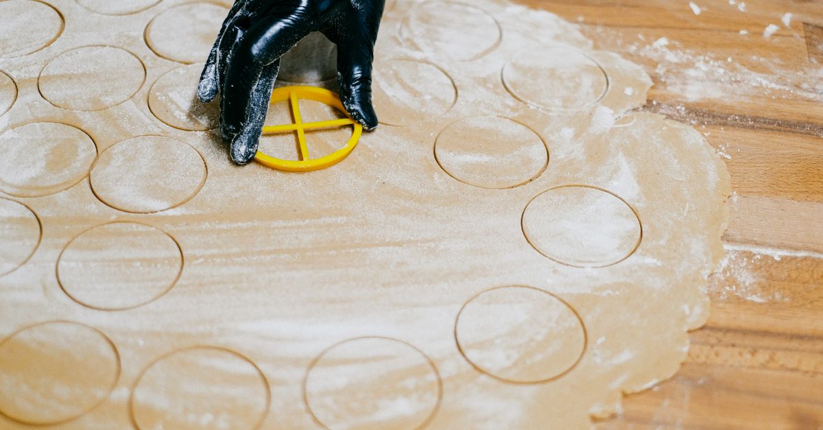 Une main gantée découpe des cercles de pâte étalée sur un plan de travail fariné à l'aide d'un emporte-pièce jaune.