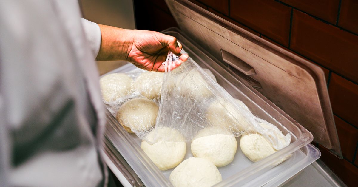 Une main recouvre d'un film plastique plusieurs boules de pâte crue disposées dans un bac en plastique.