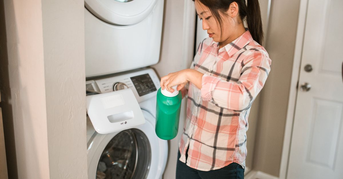 Une femme verse un produit liquide depuis une bouteille verte au-dessus d'un lave-linge domestique.