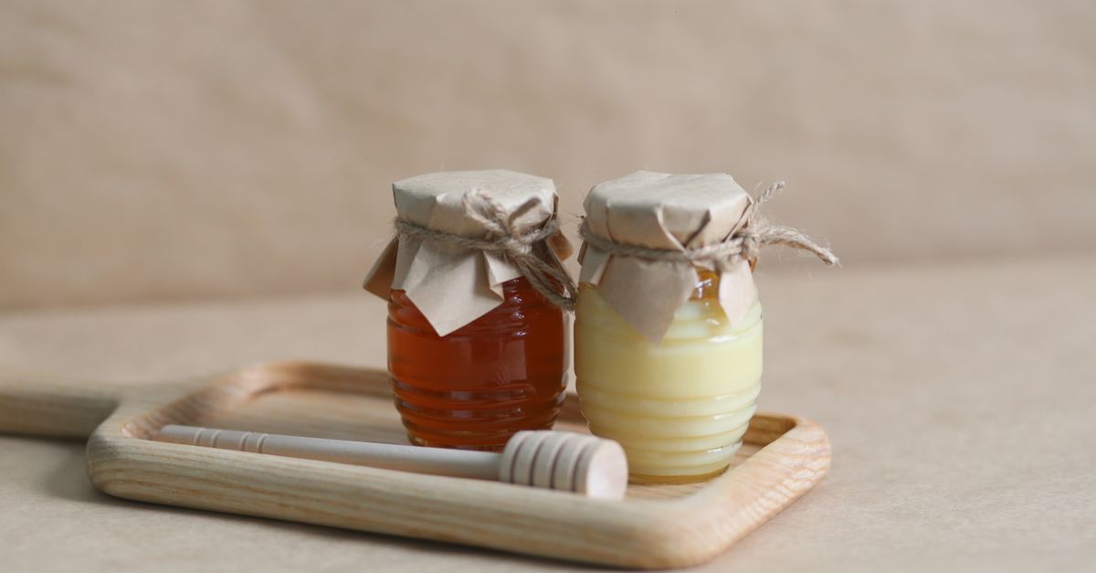 Deux petits pots en verre fermés avec du papier kraft et de la ficelle, l'un contenant du miel ambré et l'autre un liquide blanchâtre crémeux ressemblant à du lait concentré sucré, posés sur un plateau en bois avec un bâtonnet à miel.