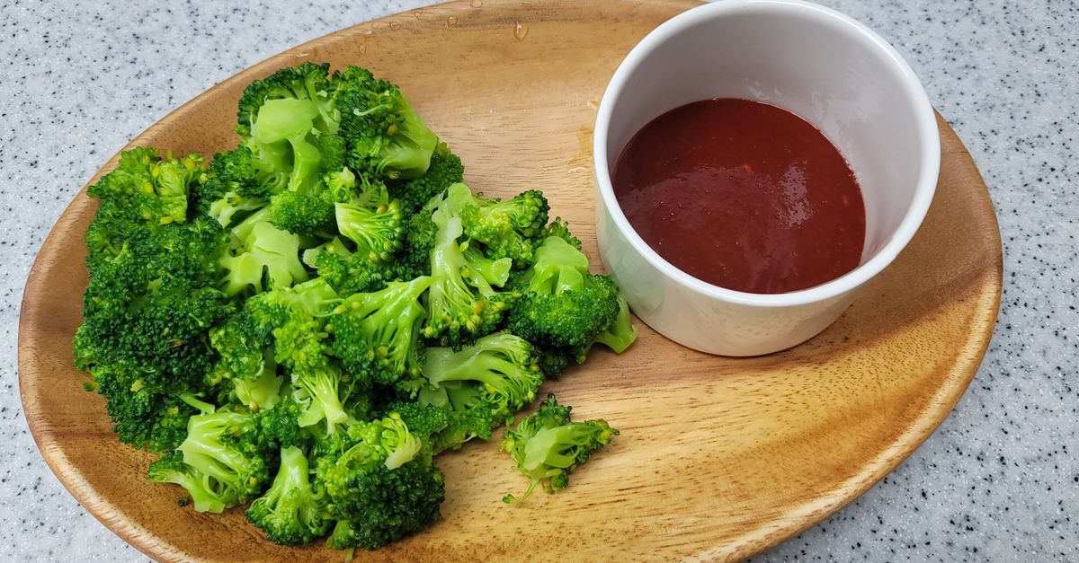 Des fleurettes de brocoli cuites disposées sur une assiette en bois, accompagnées d'une sauce rouge dans un petit bol.