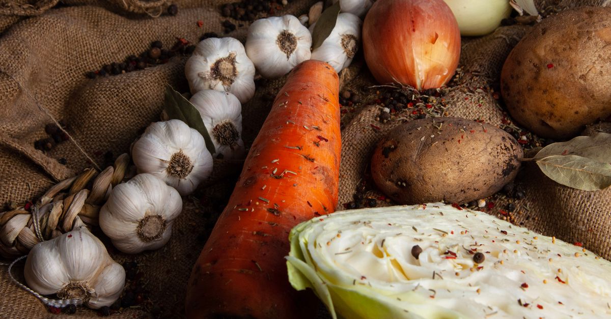 Des légumes racines d'automne (ail, carotte, oignons, pommes de terre) et un chou posés sur un tissu de jute rustique.