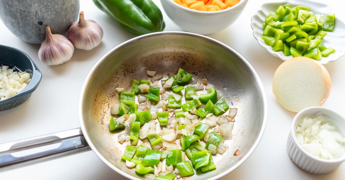 Des poivrons verts et des oignons coupés en dés cuisent dans une poêle en métal, entourés de légumes crus sur un plan de travail.