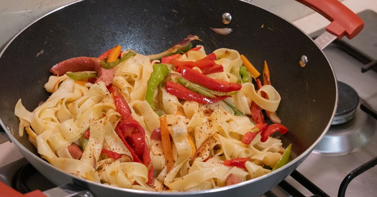 Des lanières de poivrons verts, rouges et jaunes sautées avec des tagliatelles dans une poêle sur une cuisinière à gaz.
