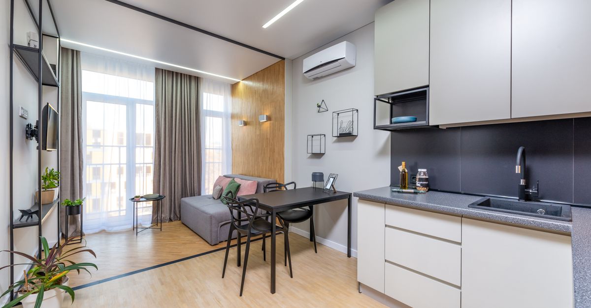 Coin cuisine compact ouvert sur un salon de studio, avec plan de travail gris, meubles blancs, plaque de cuisson intégrée et grande fenêtre apportant de la lumière naturelle.