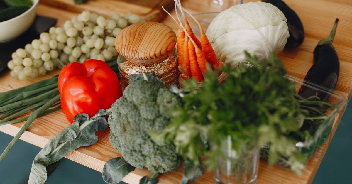 Planche en bois avec des carottes entières avec leurs fanes vertes, du brocoli, un poivron rouge et d'autres légumes disposés autour.