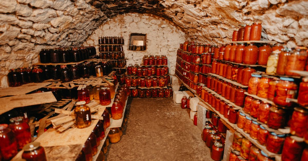 Des étagères en bois chargées de nombreux bocaux de conserves dans une cave voûtée en pierre.