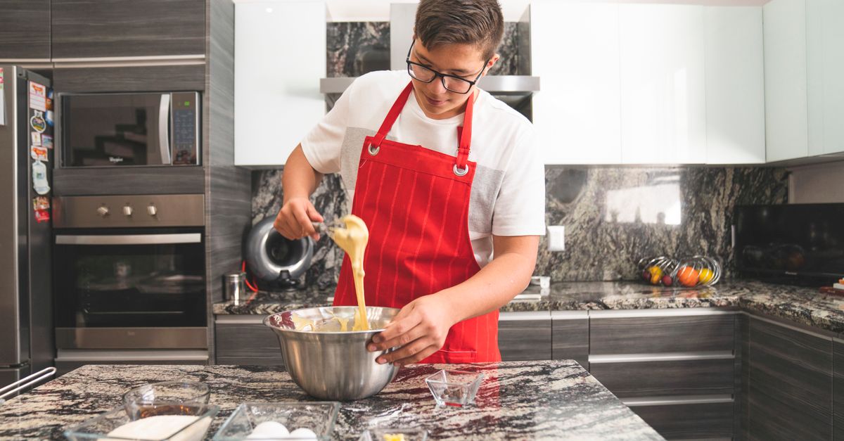 Un jeune homme en tablier rouge fouette une préparation dans un bol en inox posé sur un plan de travail en marbre, dans une cuisine moderne.