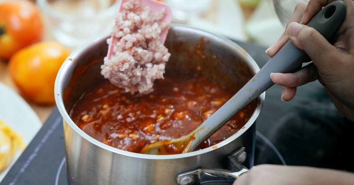 Une main ajoute de la viande hachée dans une casserole de sauce tomate en train de mijoter, remuée avec une cuillère.