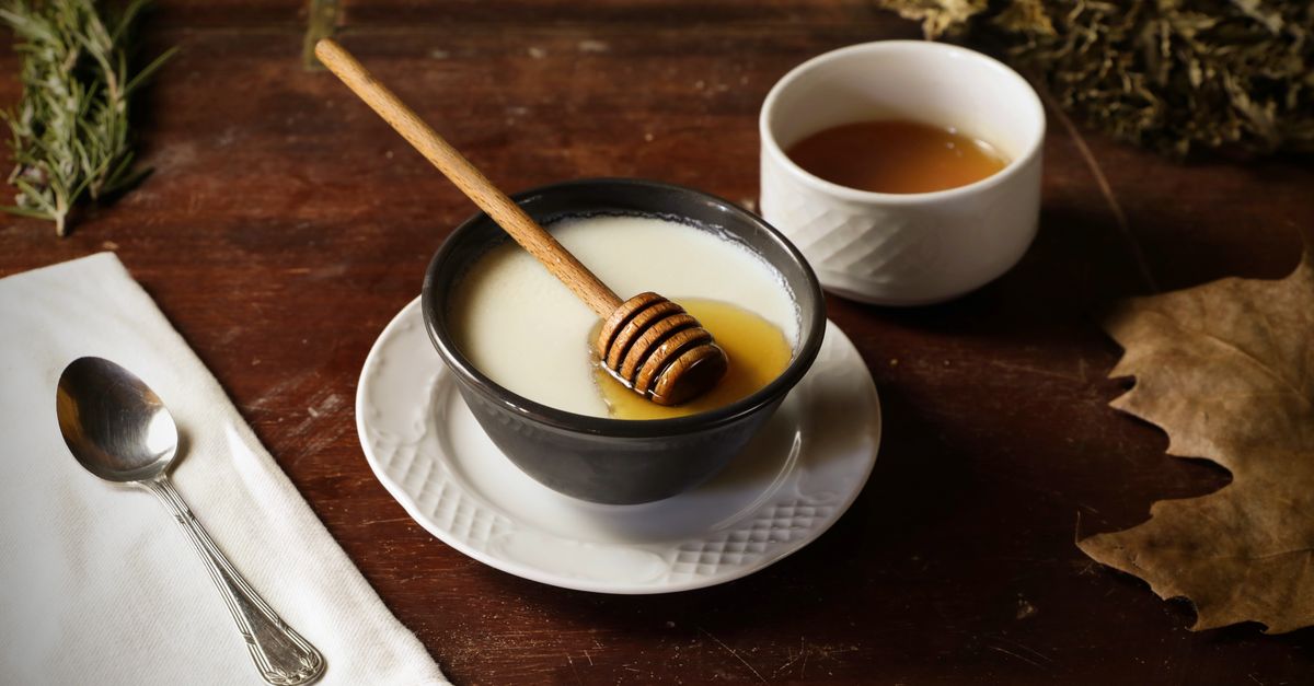Un bol en céramique rempli d'un liquide blanchâtre type beurre fondu, avec un bâtonnet à miel en bois posé dedans, à côté d'une tasse de liquide doré sur une table en bois.