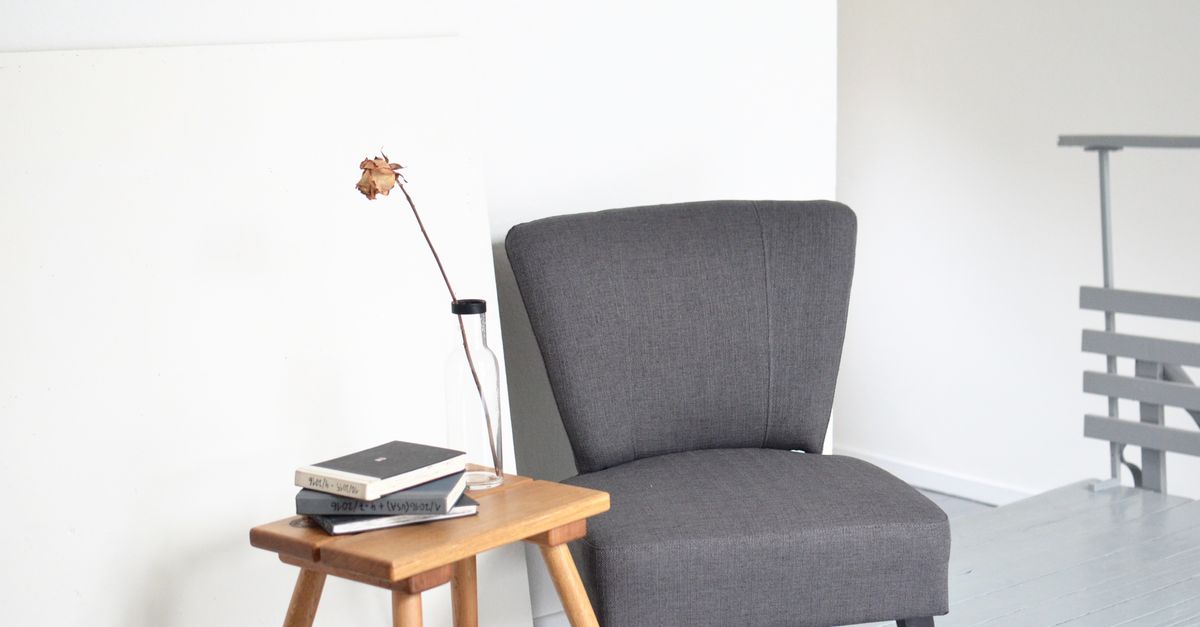 Un fauteuil gris minimaliste près d'une table basse en bois avec des livres empilés et une fleur séchée dans un vase, dans une pièce lumineuse aux murs blancs.