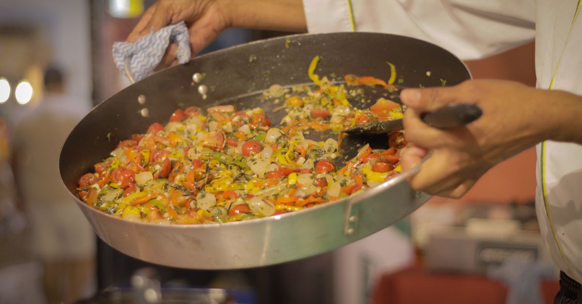 Un chef fait sauter des poivrons et tomates coupés dans une grande poêle tenue à la main.