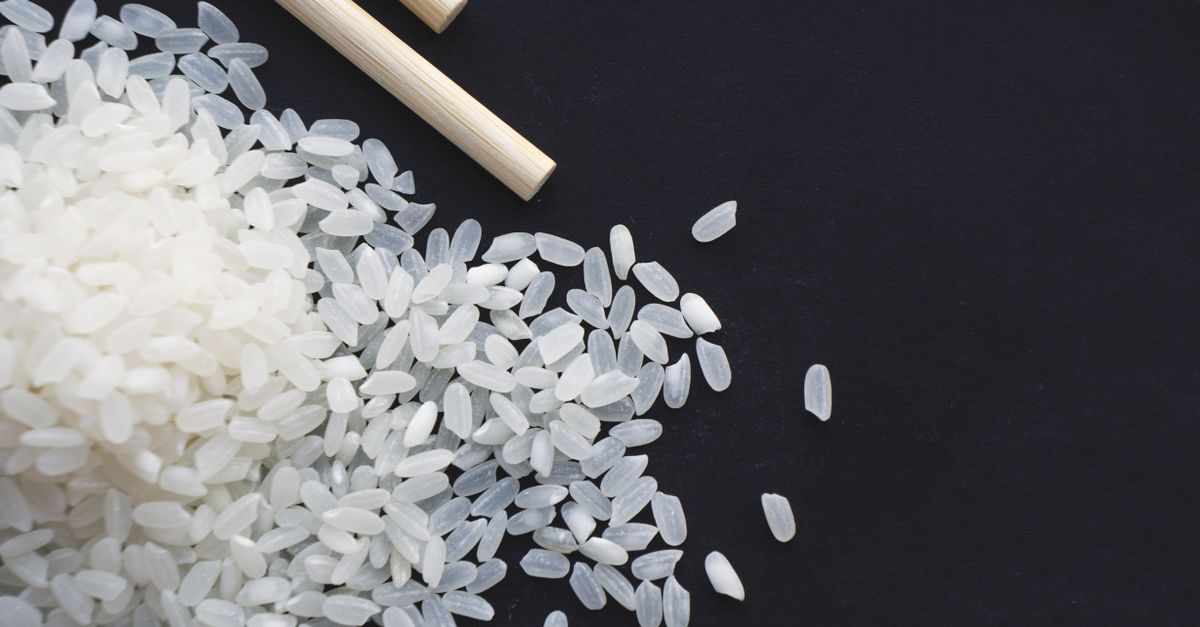 Des grains de riz blanc cru éparpillés sur une surface noire à côté de baguettes en bois.
