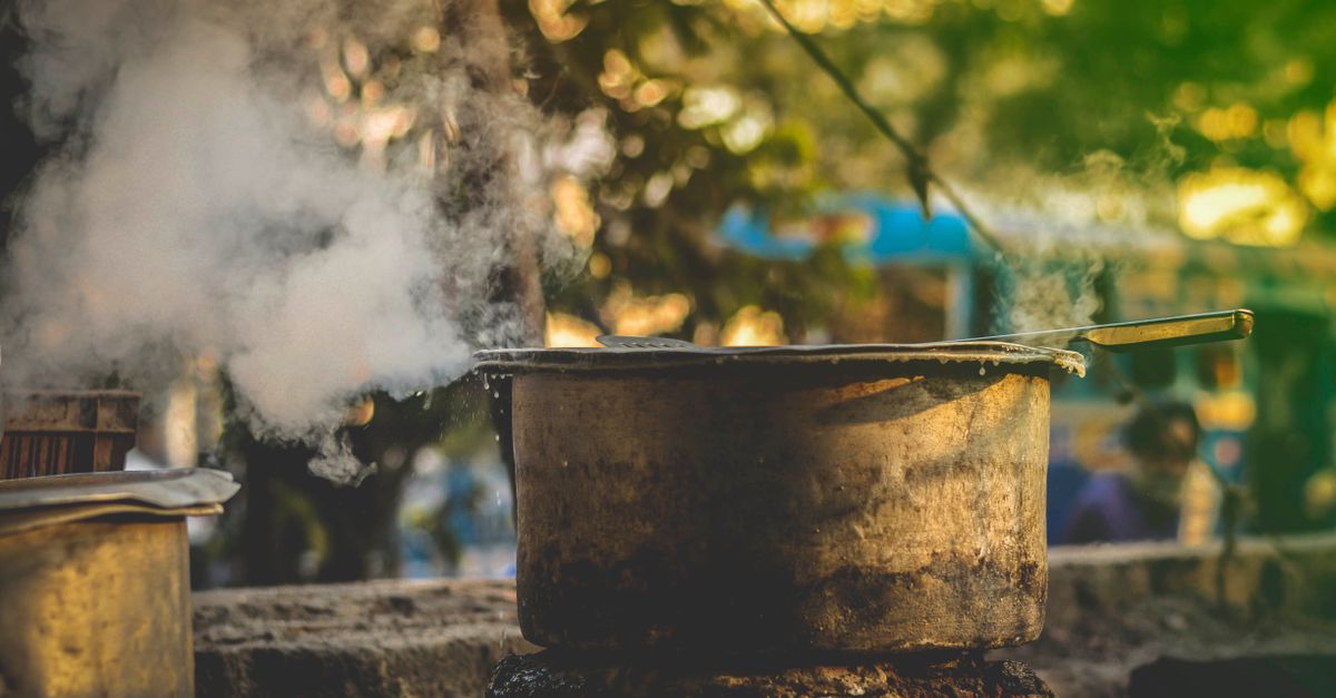 Une vieille casserole noircie fume sur un feu extérieur, de la vapeur s'échappant sous son couvercle.
