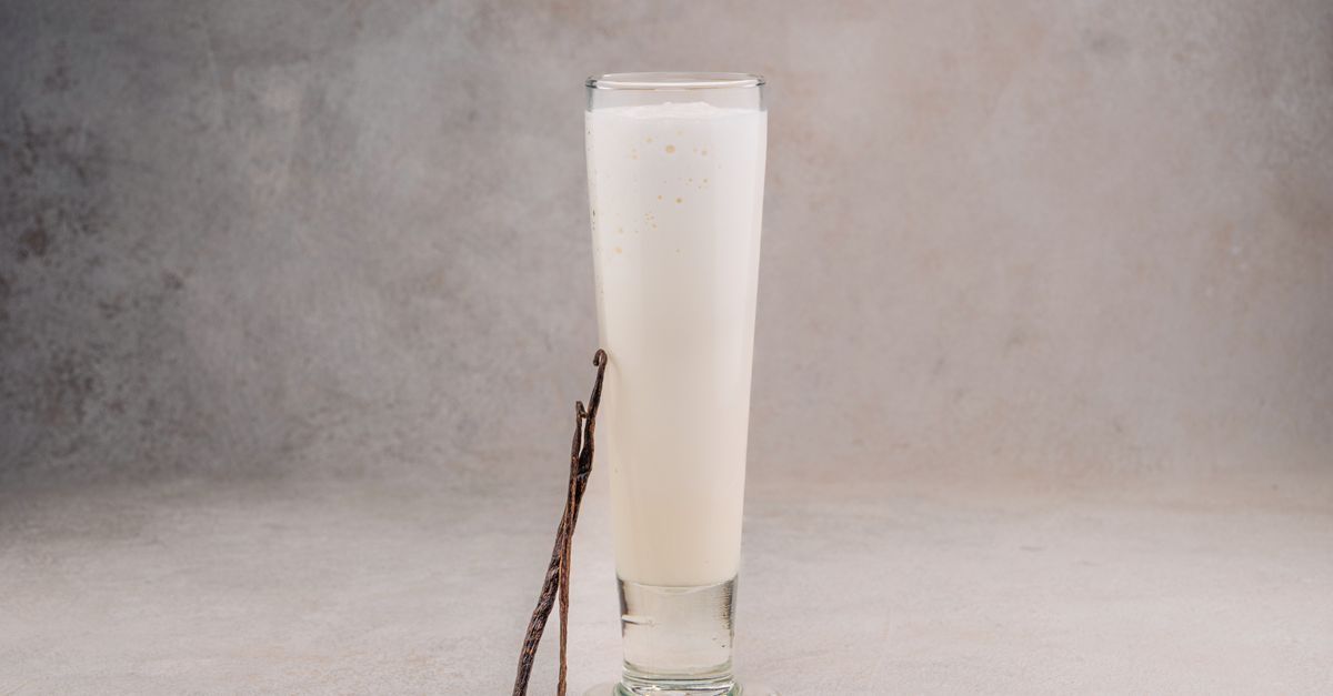 Une gousse de vanille fendue posée contre un grand verre rempli de lait, sur un fond gris minéral.